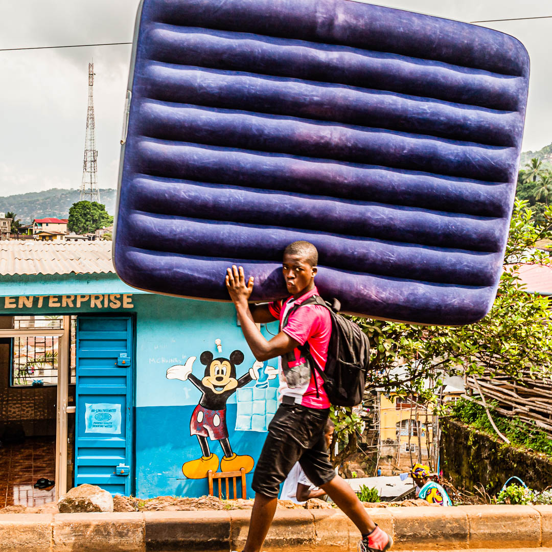 Junger Mann transportiert aufgeblasene Luftmatratze vor einem Mickey Mouse Unternehmen in der Western Area Rural, Sierra Leone / © Foto: Georg Berg