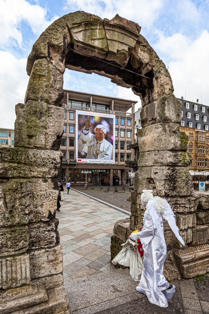Als Engel verkleideter Schauspieler am Kölner Dom vor dem Plakat von Papst Benedikt / © Foto: Georg Berg